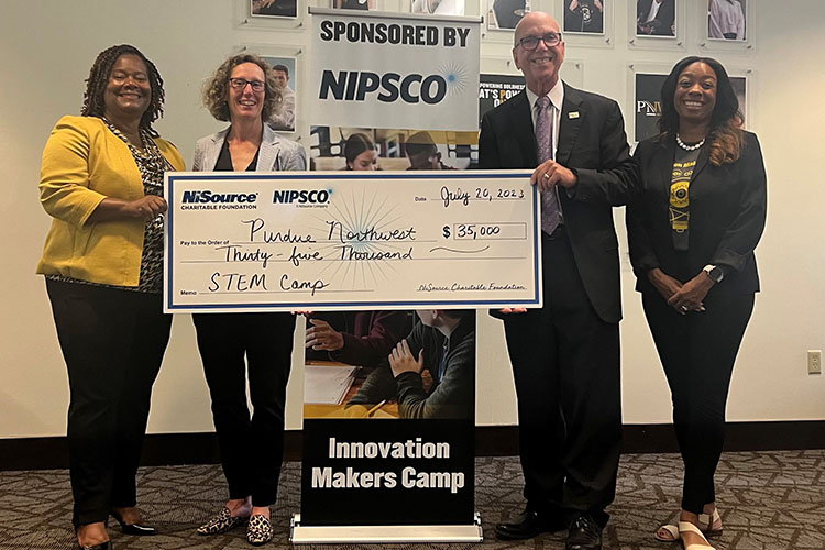 Four people pose together. Three of them are helping hold up a large check.