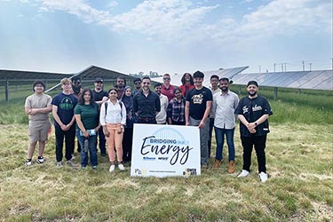 People pose in a field of solar panels with a sign that reads, "Bridging Our Energy," NiSource, Nipsco, Purdue University Northwest, Power Onward"