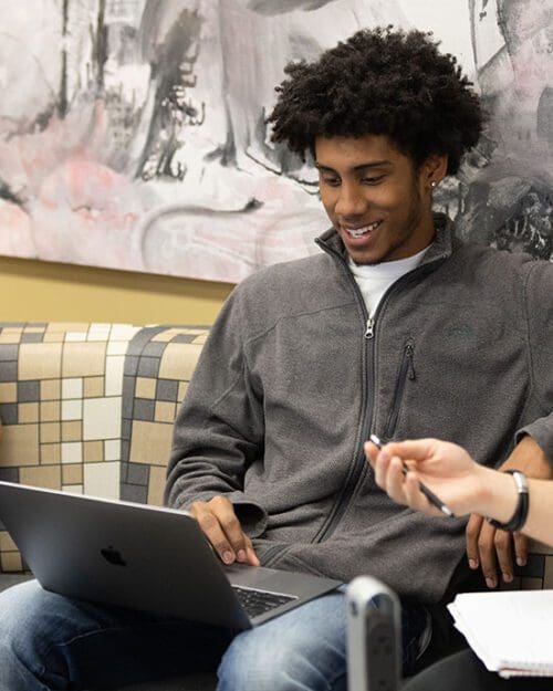 A student in a gray zip up sweater and jeans. They have a laptop open on their lap.