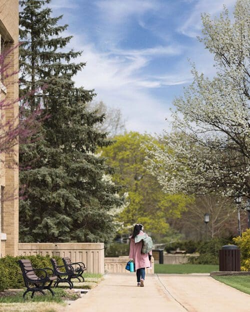 A student walks down a sidewalk on the Westville campus. They are walking away from the camera, wearing a pink coat and a backpack