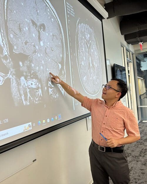 A student stands in front of a projector screen. Their body is facing the camera while their head is looking at the screen. They are wearing a light orange shirt and gray pants. There is an MRI on the screen and the student is pointing to part of it.