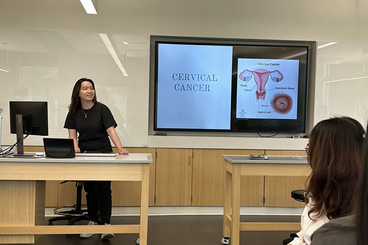 A student in a black shirt and pants stands behind a desk while giving a presentation. The screen behind them reads "Cervical Cancer"