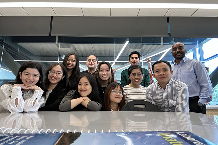 11 people pose together. The front row of people are leaning on a table, the second and third row are leaning in a little bit.