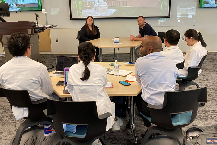 Five students in lab coats sit at a table. Their backs are facing the camera. There are two Biological Sciences staff members sitting at a different table and facing the camera.