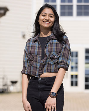 A student in a brown and blue flannel and black jeans stands in front of a university village building