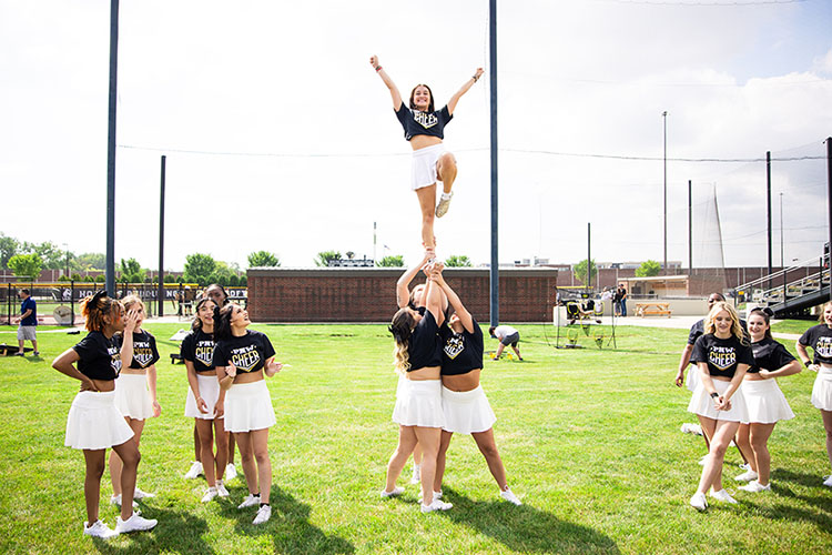 A PNW cheerleader stands and smiles while being lifted into a pyramid. Other cheerleaders smile and watch