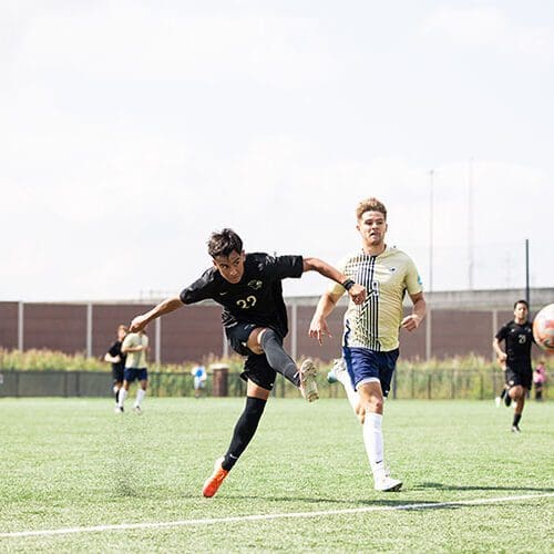 Two men's soccer players run down the field. The ball is pictured mid-air
