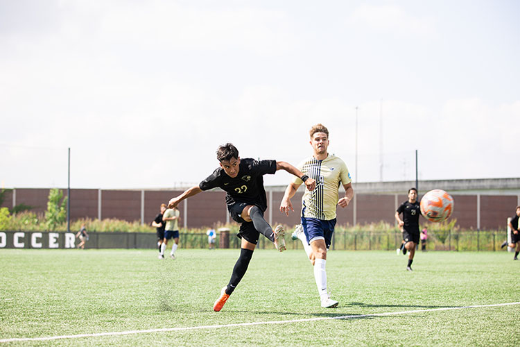 Two men's soccer players run down the field. The ball is pictured mid-air