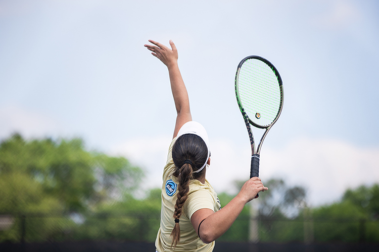 A PNW womens tennis player throws a ball into the air to serve. She is holding a tennis racket.