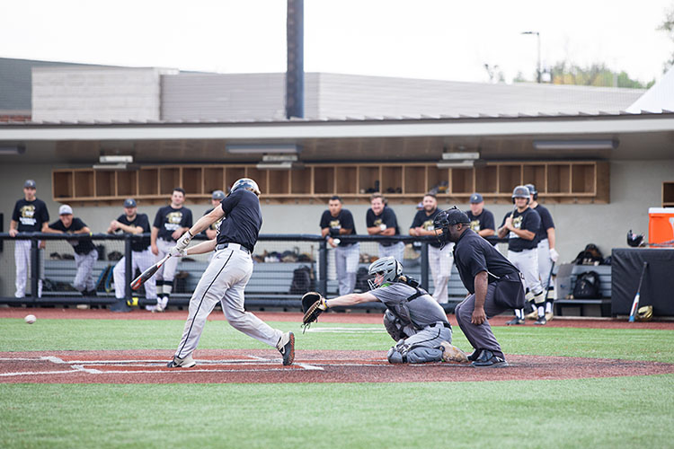 A baseball player swings at a ball. The catcher has his arm extended. The PNW baseball team can be seen standing in the dugout in the background.