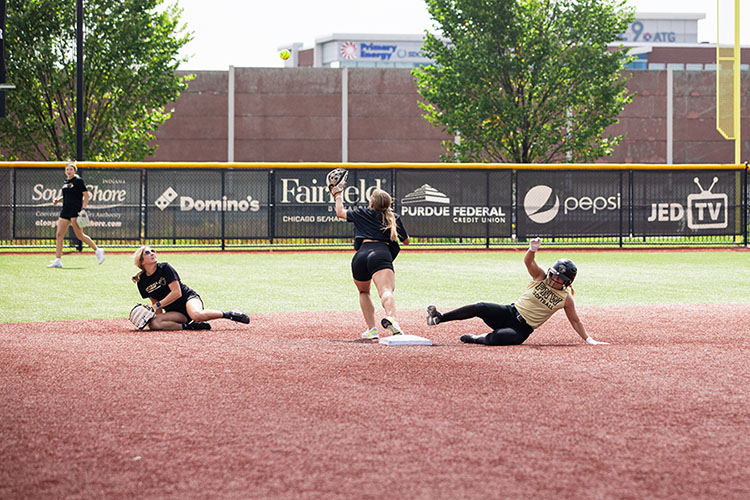 A PNW softball player slides into second base. Another player is outstretched on the base to try and catch the ball. A third player is laying on her side in the dirt.