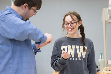 A student smiles while wearing a PNW sweatshirt. Another student stands in a blue shirt and is slightly turned away from the camera.