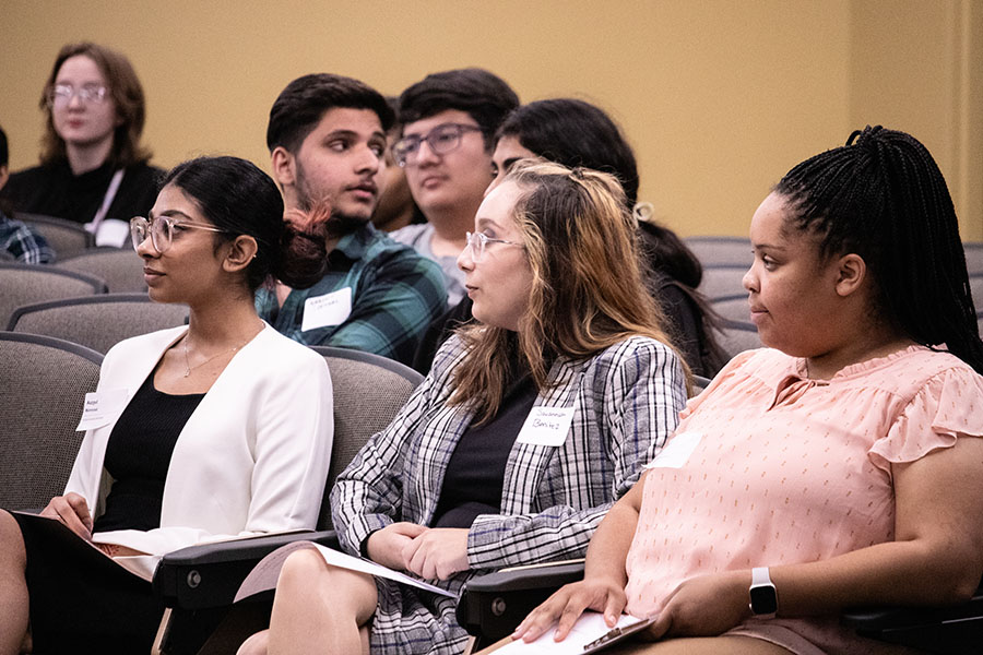 Students sit in chairs, looking at a stage.