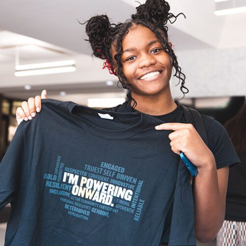 A student smiles and holds up an "I'm Powering Onward" t-shirt