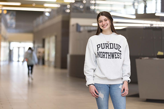 A student stands in a gray Purdue Northwest sweatshirt and light jeans.