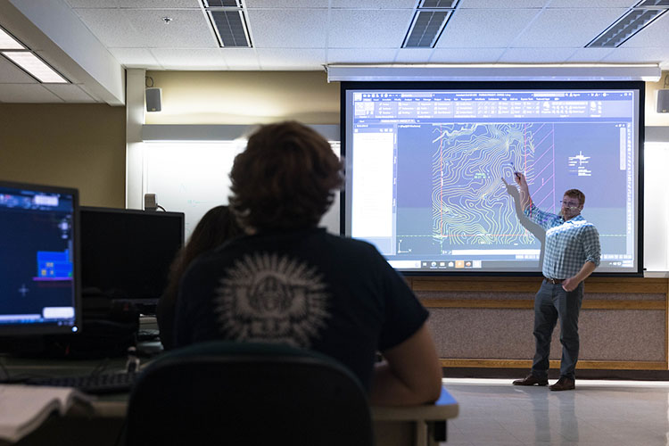Students sit in a classroom and look at a professor. The professor is pointing to a part of a map that is being projected onto the screen behind them.