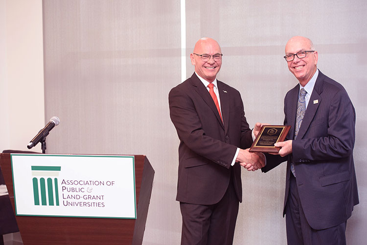 Two men shake hands and pose with a plaque near a podium reading "Association of Public and Land-Grant Universities."