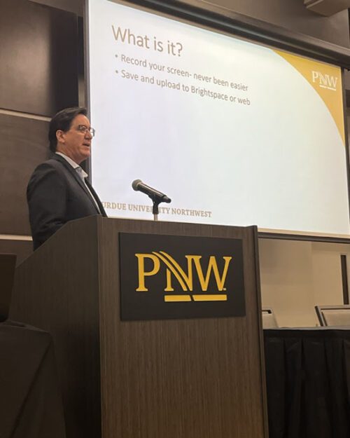 David Pratt stands behind a wooden podium with the "PNW" logo on it. He is speaking into a microphone. A slideshow for his presentation is being projected onto the screen behind him