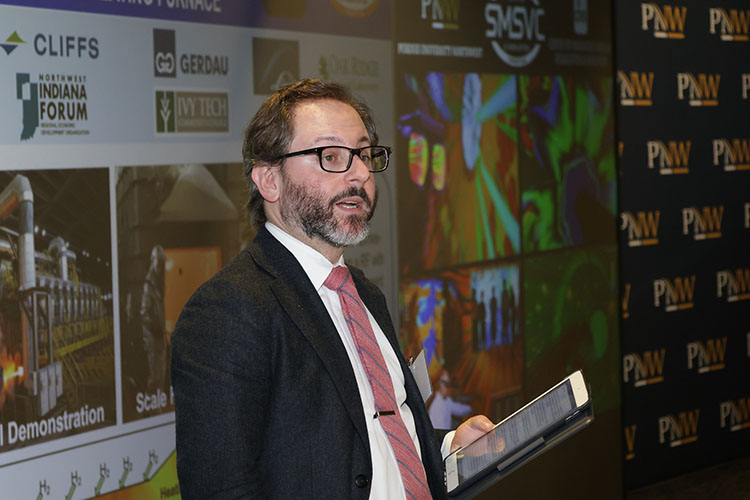 Avi Shultz holds a tablet on a stage and speaks to a crowd