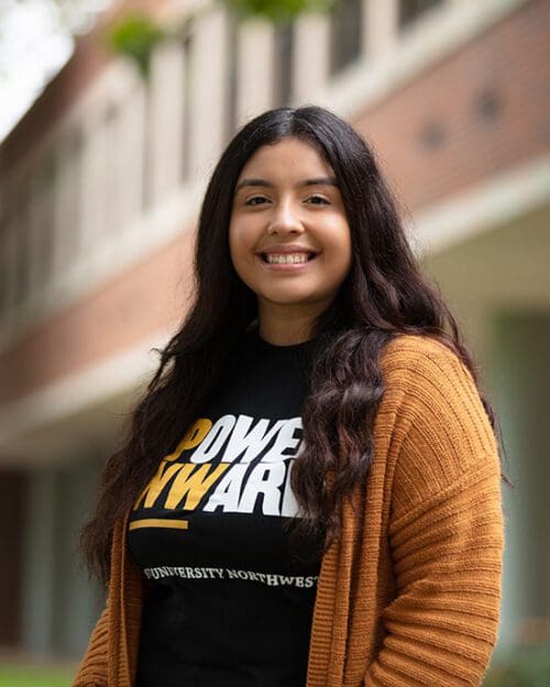 A student looks at the camera and smiles. They are wearing a black Power Onward t-shirt and an orange-brown cardigan