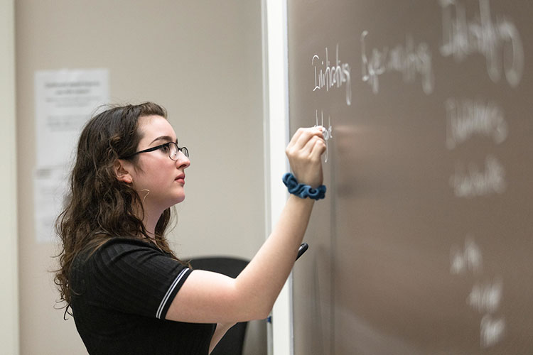 A student in a black t-shirt writes on a chalk board