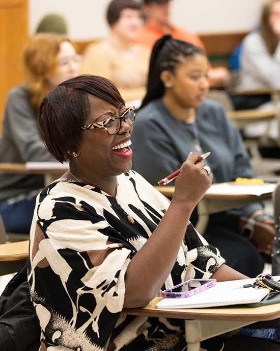 A student sits in desk in a classroom. Their elbow is propped on the desk and they are holding a pen in the air.