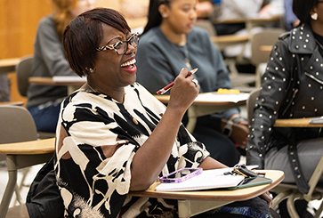 A student sits in desk in a classroom. Their elbow is propped on the desk and they are holding a pen in the air.