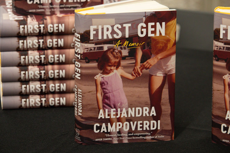 The book, "First Gen: A Memoir," stands upright to present its cover with an older image of a child whose hand is held by an adult. A stack of the same book lies in the back.