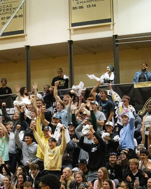 Crowd of students sit on indoor gymnasium bleachers as they cheer on athletics event.
