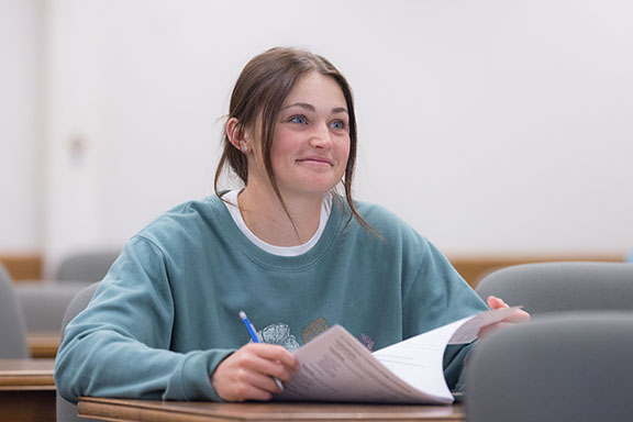 A student sits in a classroom. They are wearing a blue sweatshirt and holding papers.