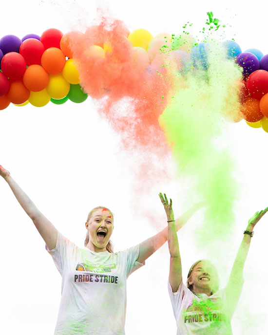 Two students stand under a rainbow balloon arch and throw green and orange color powder in the air