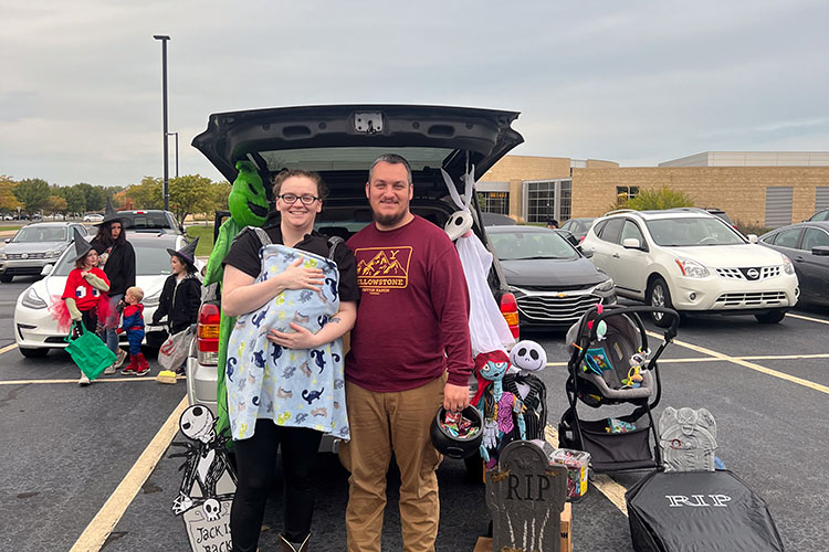 Man and woman pose with their Halloween-decorated trunk with a bowl of candy.