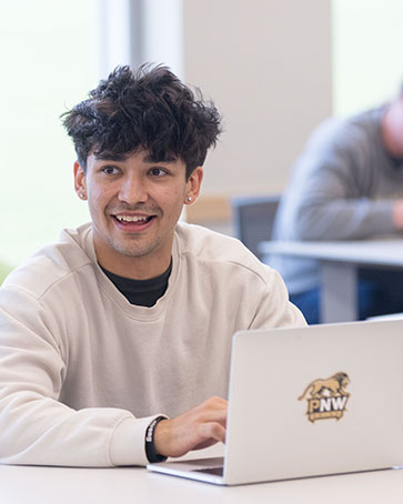 A student in an off-white sweatshirt sits at a table. There is a laptop open on the table.