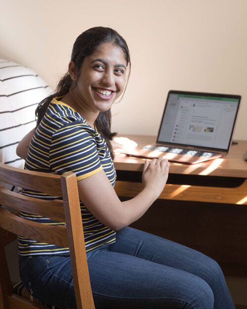 A student sits at a desk in their dorm room. They are wearing a black and white striped shirt and blue jeans. Their hands are resting on an open laptop.