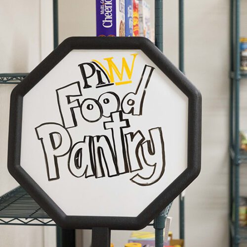 An octogon white board sign that reads "PNW Food Pantry" The food pantry shelves are in the background.