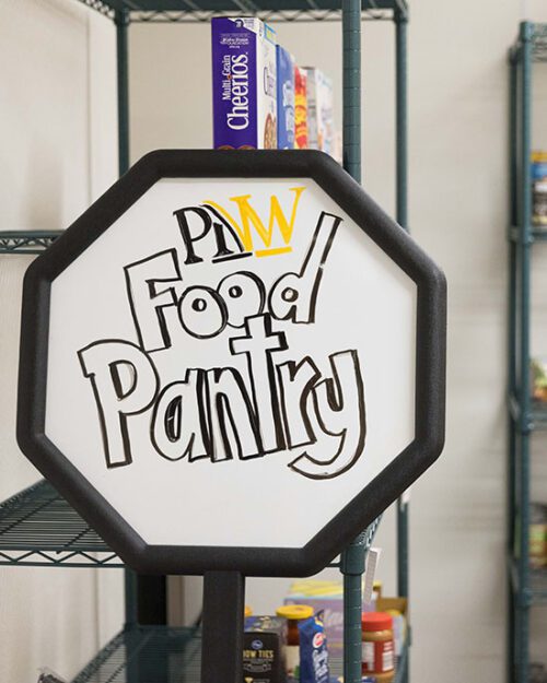An octogon white board sign that reads "PNW Food Pantry" The food pantry shelves are in the background.