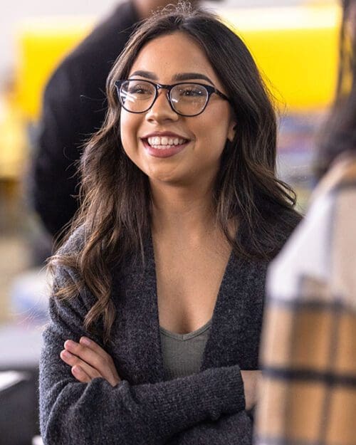 A student in a light gray shirt and dark gray cardigan stands with their arms folded. They are looking off to the left and smiling.