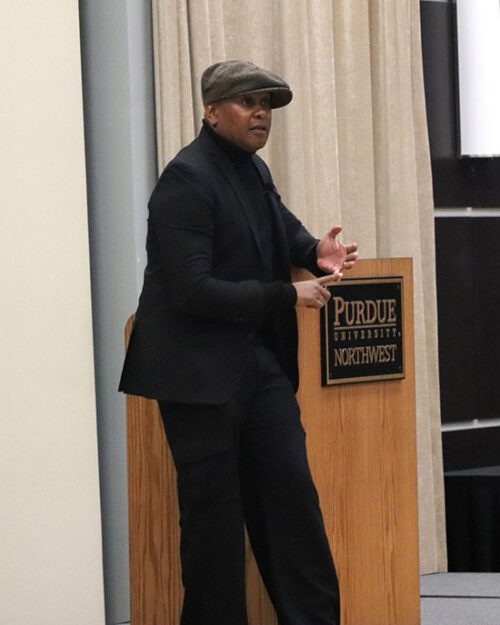 Kevin Powell stands on stage wearing a black quit and a gray hat. He is leaning against a podium