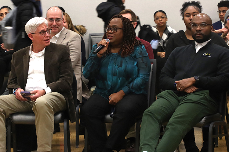 Students and staff sit in rows of chairs. La Tonia Winston is in the center of the photo, sitting down and holding a microphone.