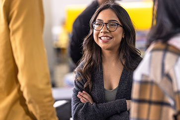 A student in a light gray shirt and dark gray cardigan stands with their arms folded. They are looking off to the left and smiling.