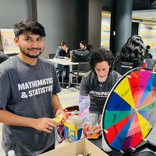 A student in a "Mathematics & Statistics" stands next to a prize wheel.