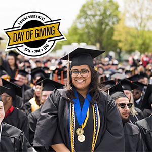 A student in commencement regalia poses next to a badge that says Purdue Northwest Day of Giving 04/24/24