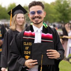 A Spring 2024 graduate in a Latino affinity stole and commencement regalia holds their cap in front of them and smiles
