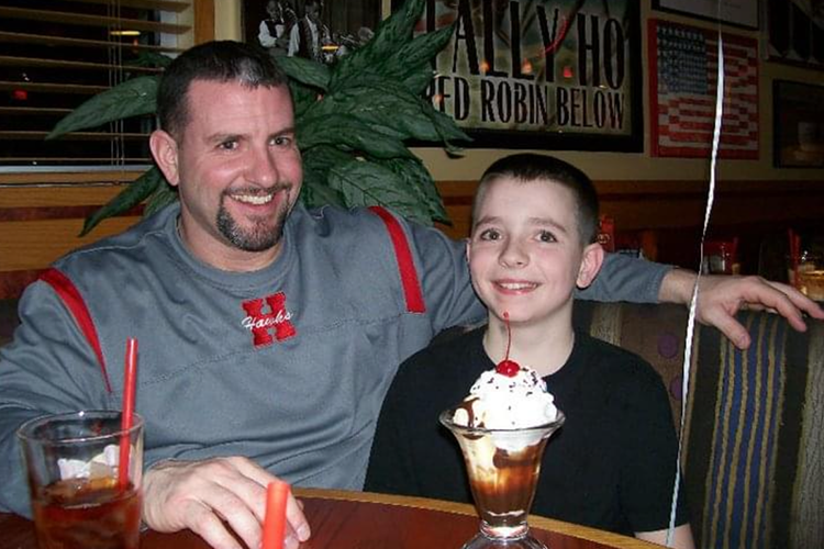 Jacob Wheeler Sr. and Jacob Wheeler Jr. sit next to each other in a booth when Jr. was still a child. There are ice cream sundaes in front of them.