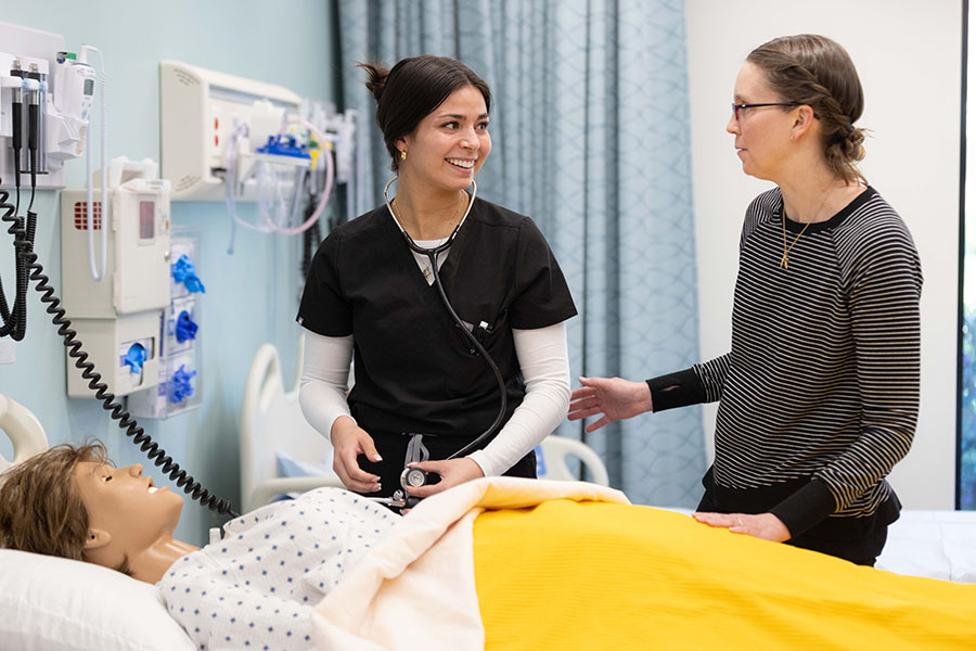 Nursing student wears stethoscope to treat a dummy-patient while talking to instructor