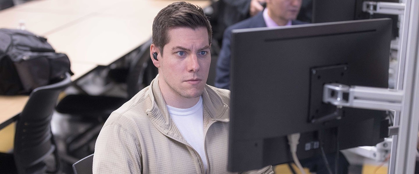 A student works at a computer in a classroom. They are wearing a beige quarter zip and a white t-shirt.