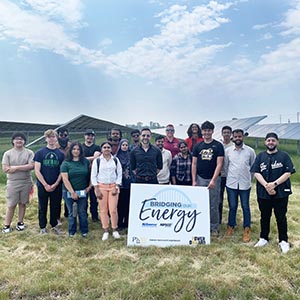 People pose in a field of solar panels with a sign that reads, "Bridging Our Energy," NiSource, Nipsco, Purdue University Northwest, Power Onward"