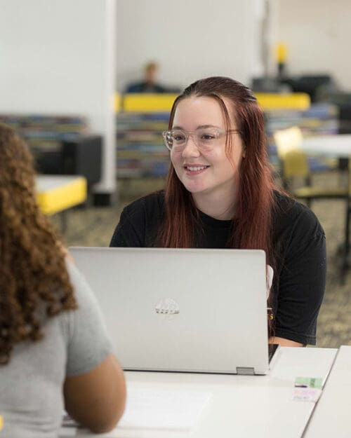 A student sits at a table with a laptop open in front of them. They are looking at another student off camera.