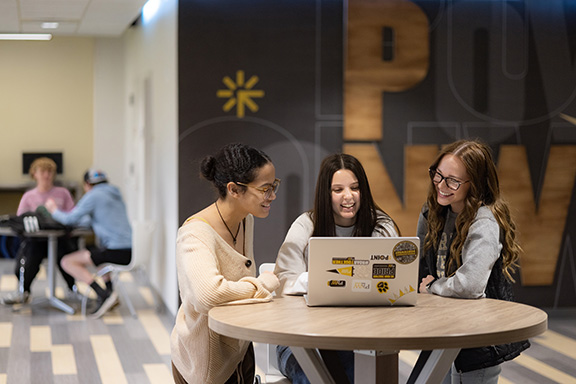 Three students sit around a circle table. They are working on a laptop that has PNW branded stickers on it.