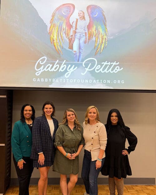 Gisele Casanova, Maureen Bauer, Nicole Schmidt, Tara Petito and Nicky Ali Jackson pose together under a projector screen that displays the logo for the Gabby Petito Foundation.